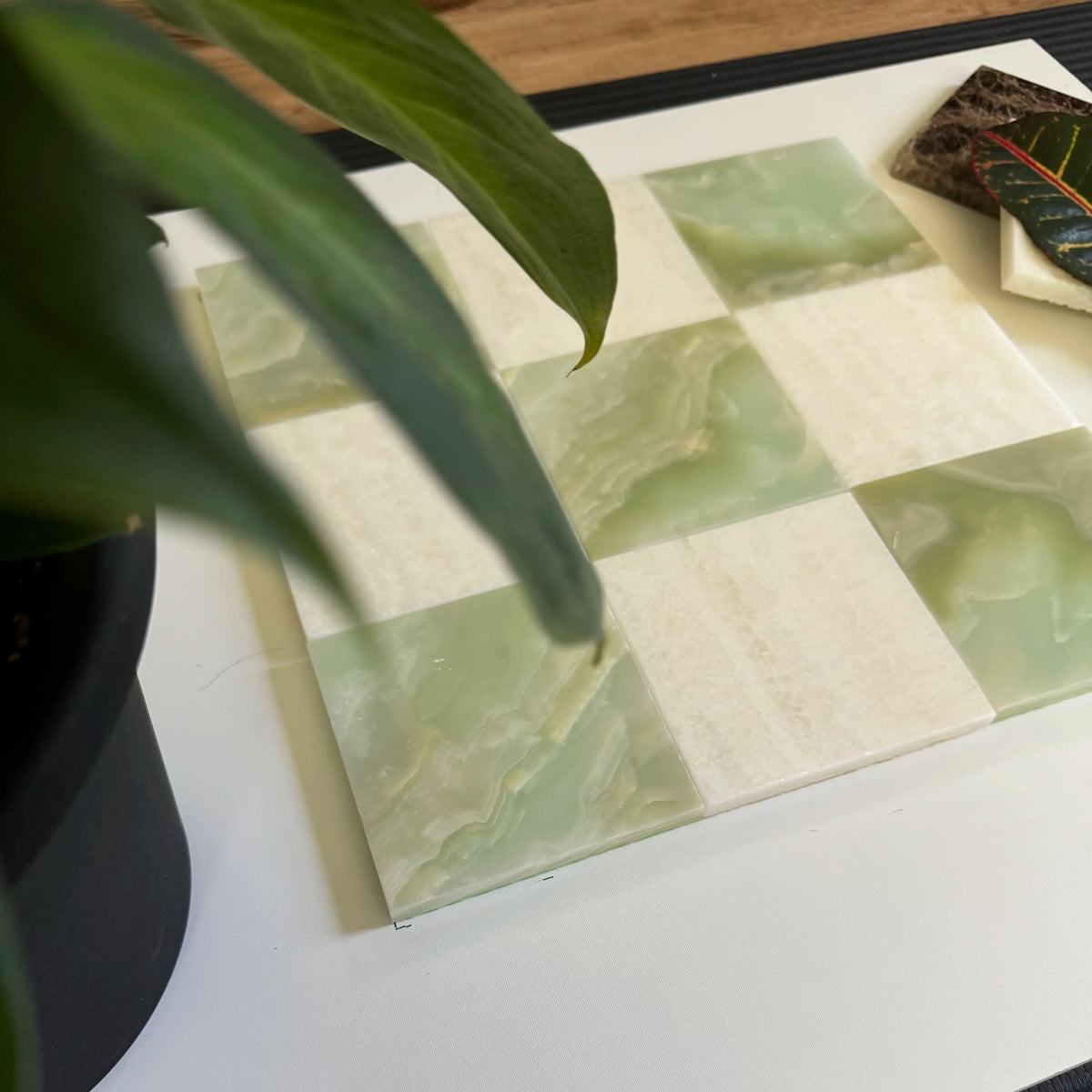 Green Onyx and Vanilla Onyx tiles arranged in a checkerboard pattern, framed by lush green leaves. The smooth surface and earthy color palette of these onyx tiles bring a calming effect, suitable for accent walls or shower tile designs in high-end projects.